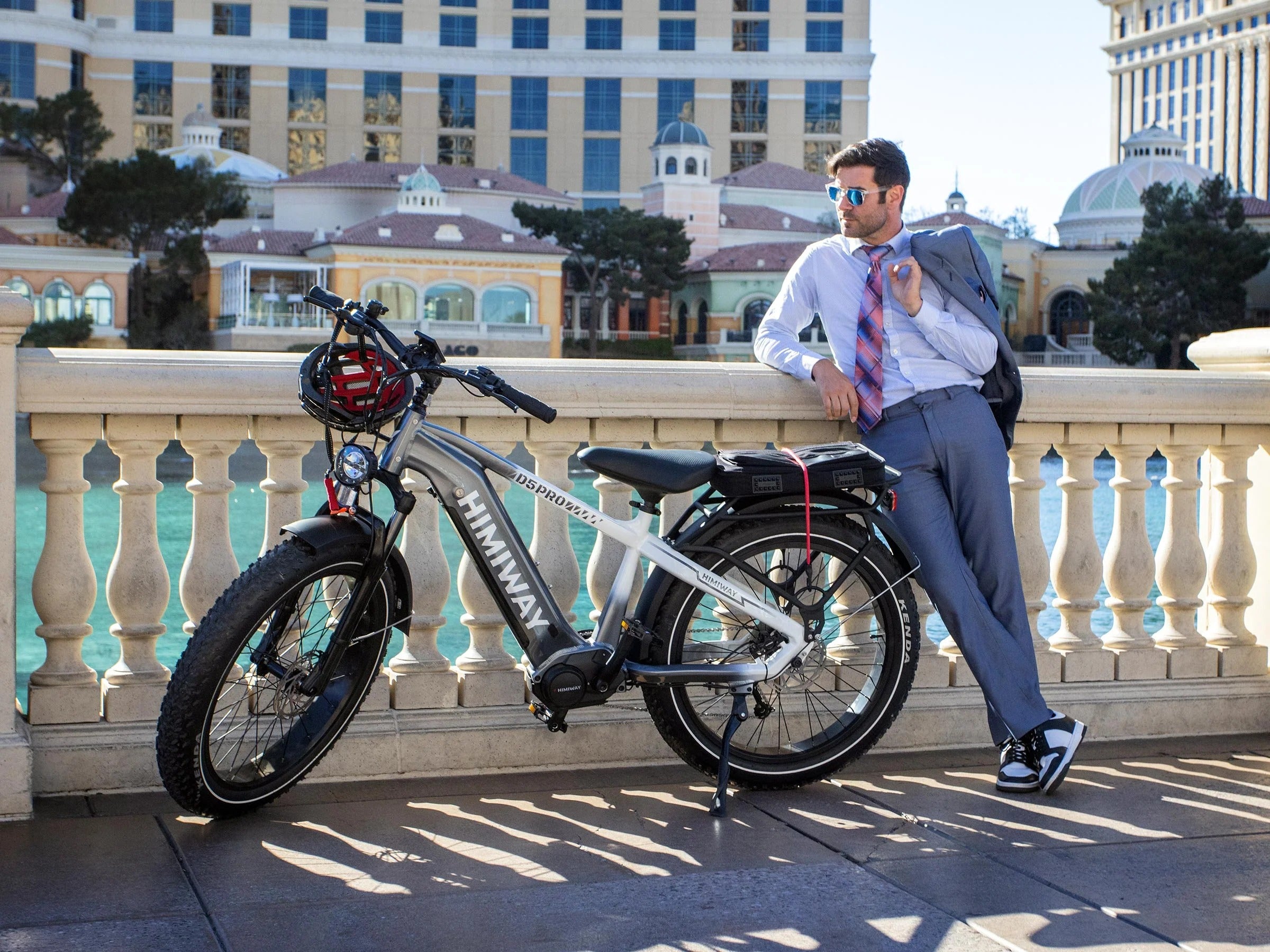 Man in a suit leaning against a railing with an electric bike in front of a scenic background