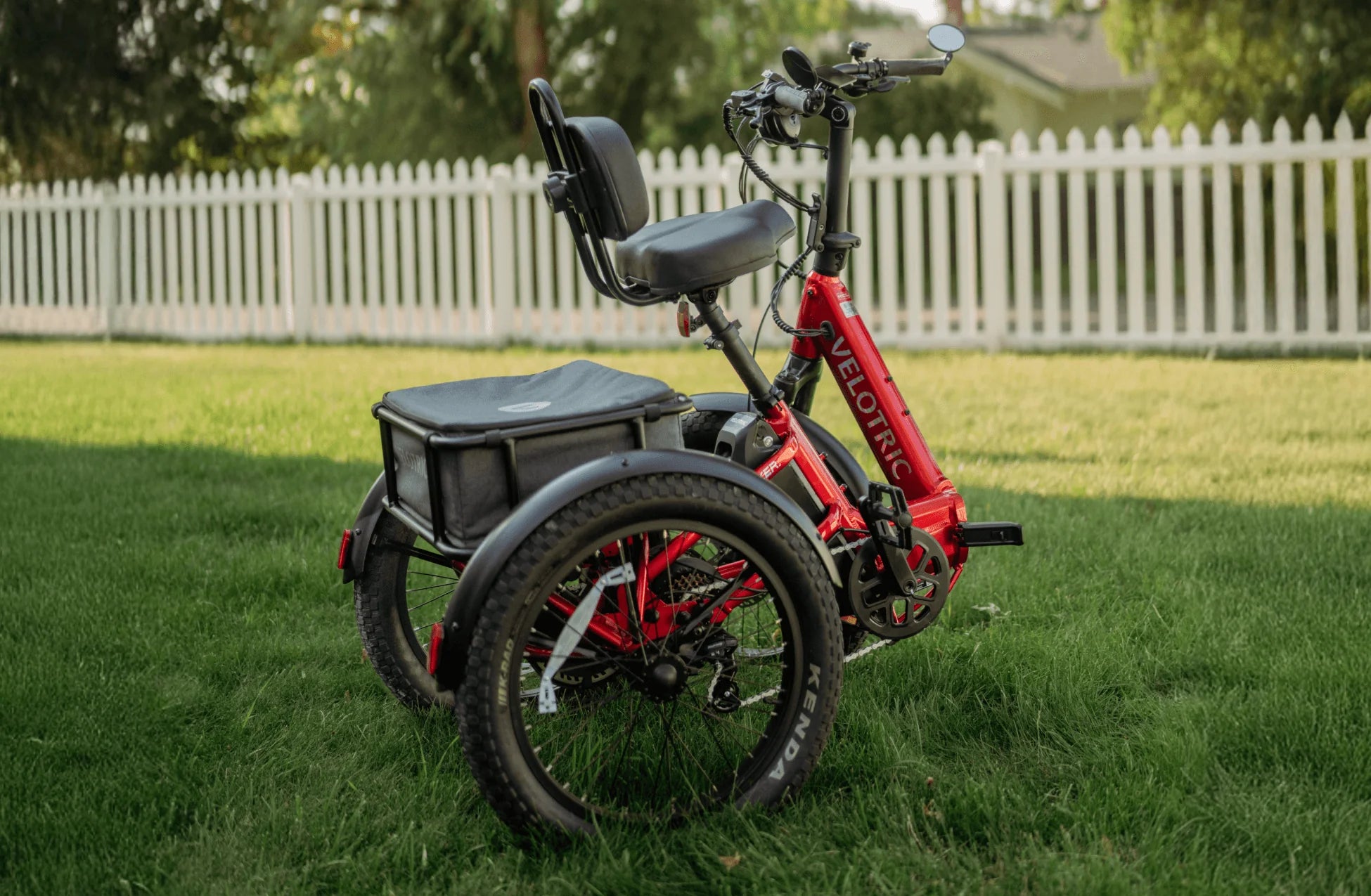 Red electric tricycle with a black seat and storage box on grass, white picket fence in background