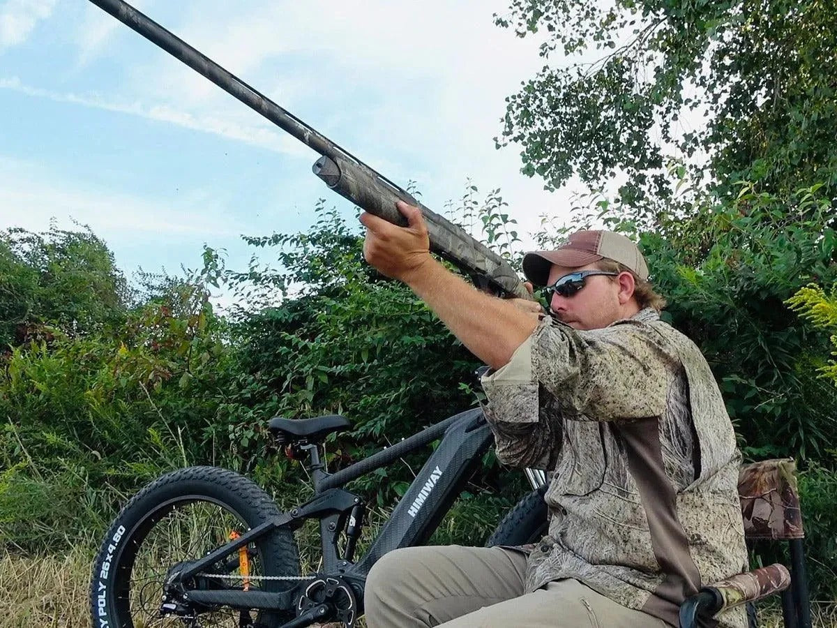 Person in camouflage gear holding a shotgun, sitting on a bike in a natural setting